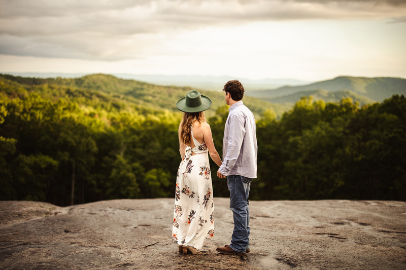 Wedding Photographer, a man and woman, husband and wife to be, hold hands as they stand on rocks staring down at the forests below them