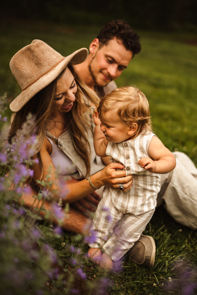 Family Photographer, mom and dad sit and admire baby in a grassy field